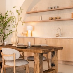 Sfeervol modern interieur met een robuuste houten eettafel, vier lichtgrijze eetkamerstoelen op een vloerkleed, en een nis in de muur met een houten keukenblok en wandplanken.